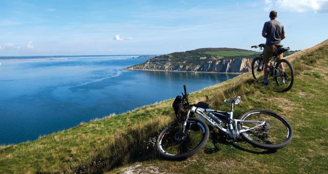 One bike lying on the ground with another being sat on as a man looks out from a clip top over the sea