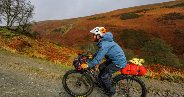 A man rides a mountain bike on a hilly gravel path, his bike weighed down by bikepacking bags. He is wearing full waterproofs and a helmet