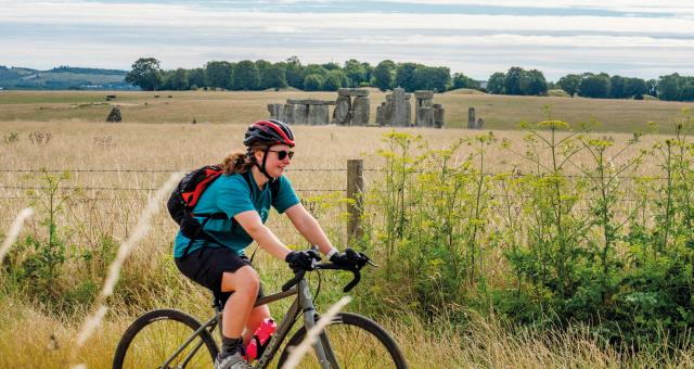 A female cyclists rides past a field in summer with Stonehenge in the background 