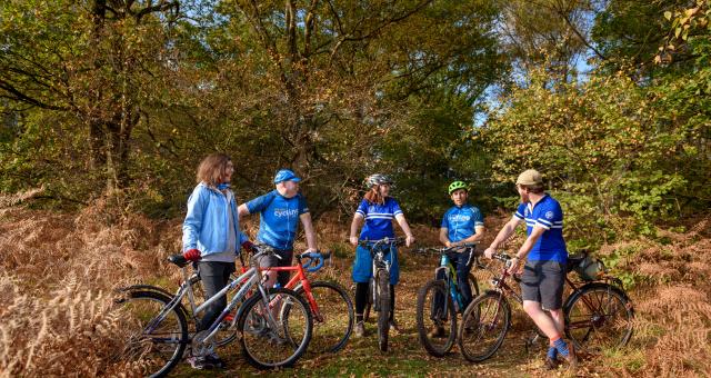 Five cyclists standing on their bikes talking to each other while in a field with trees behind them