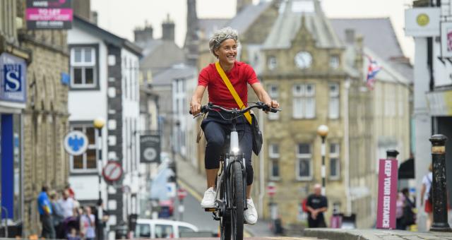 A woman is riding along a town centre road on an e-cycle. She is wearing a red T-shirt and black three-quarter length trousers with a messenger bag across her body. There are shops and businesses in the background