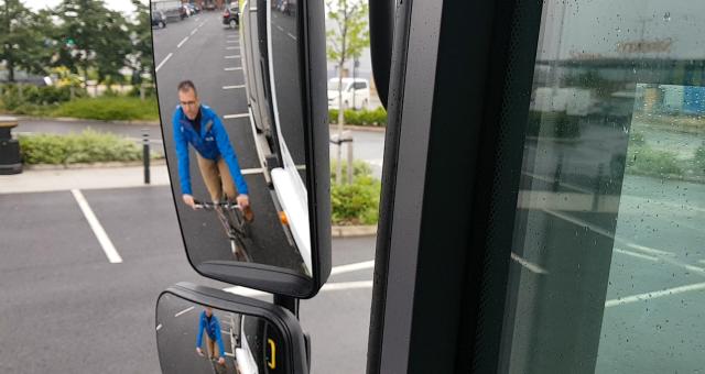 A close-up of a left-hand-side wing mirror on a lorry showing a man on a bike in the background