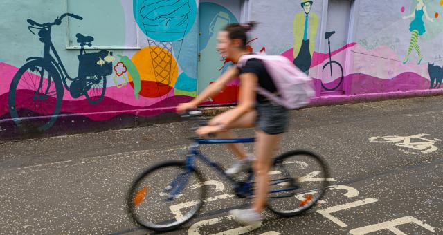 A woman is cycling on a blue bike. She's wearing shorts and T-shirt and has a pink rucksack. She's deliberately blurred so the background is in focus. It shows a colourful mural with bikes and ice cream and people