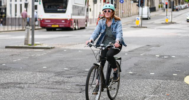 A woman is riding a black hybrid bike with a rear rack on it on an urban street. She is wearing black trousers, a stripy top and blue denim jacket. She has long red hair, a turquoise cycling helmet and sunglasses.