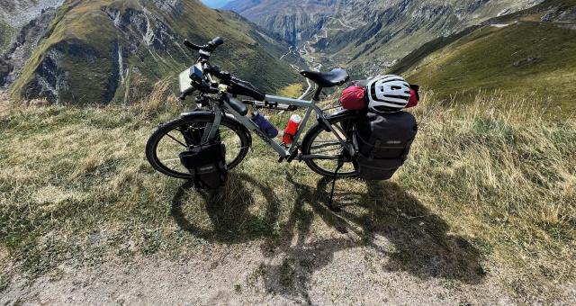 A loaded flatbar touring bike with front and rear panniers, saddlebag and top tube bag is propped up at the top of a mountain with a valley laid out behind it