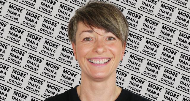 A head and shoulders shot of a woman standing in front of a screen with the More Than a Cyclist logo printed all over it. She is wearing a black T-shirt with the same logo in white. She has short hair and is smiling at the camera.