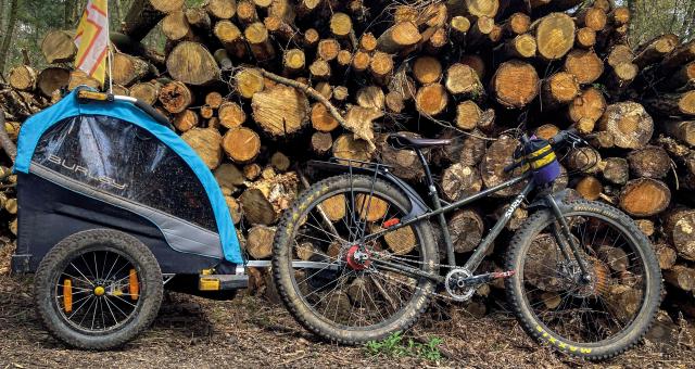 A black Surly mountain bike with a blue and black child trailer attached is leaning against a big log pile in a forest