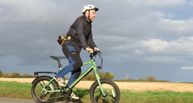 A man is riding a green e-bike with 20in wheels. He's wearing jeans, trainers, a jacket and cycling helmet. He's standing out of the saddle.