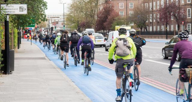 Lots of cyclists are riding along a blue segregated cycle lane next to a busy urban road. They are on a variety of bikes and wearing all kinds of clothing. Several have backpacks and it looks like they're cycling to work