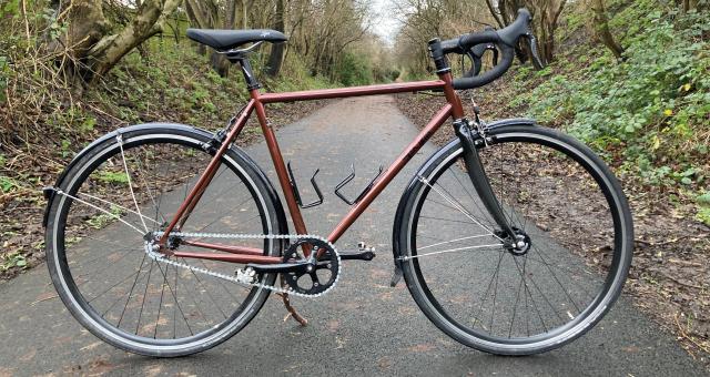 The Spa Cycles Audax Mono, a bronze fixed-wheel bike, propped up on a tarmac tree-lined path 