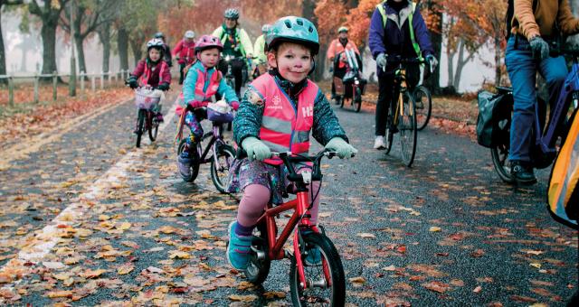 Group of cyclists including children cycling on the road, close up of a little boy on his bike wearing a helmet and high vis jacket