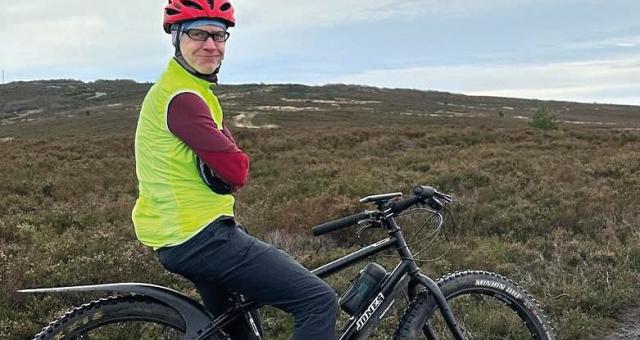 A man is sitting astride a mountain bike on a muddy path. He's got his arms crossed and is smiling at the camera. He's wearing MTB trousers, a red helmet and jersey and a bright yellow gilet.