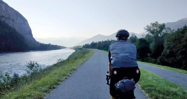 A man is cycling away from the camera on a paved cycle path alongside a river, with mountains in the background. He's wearing cycling kit and riding a touring bike with a packed seatpack