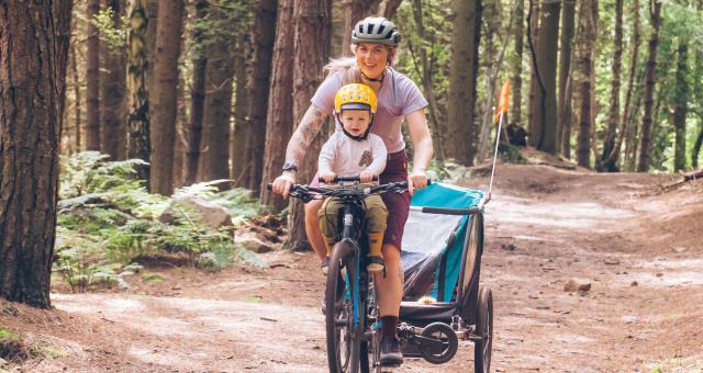 Kelly-Jayne Collinge is riding her bike on a woodland trail. Her with son Atlas is sat up front and she is pulling a trailer behind her. Mother and son are both wearing pink tops and helmets and both are smiling