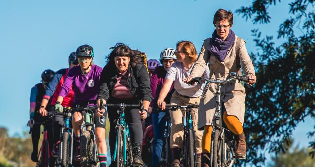 A big group of women is cycling on a tree-lined cycle path. They are wearing normal clothes and on a mix of bikes. They are smiling and chatting