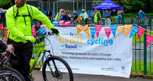 A man stands stationary with a bicycle in a park in front of a banner which says the Big Bike Revival.