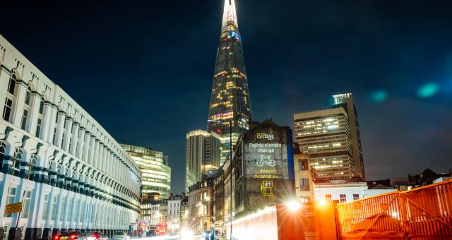 Projection in front of 'London's pyramid', the Shard building, with the words "Fights climate change" above a picture of a bike and "doesn't depend on" above a petrol pump