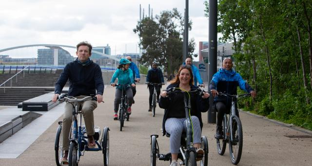 A group of people are cycling along a paved off-road path. They are on a mix of bikes but all are e-bikes.