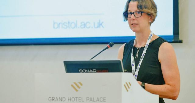 Fiona Spotswood behind a lectern with a laptop and mic propped on it. She is giving a lecture at a university.