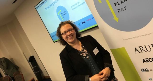 Helen Hemstock is wearing a black blazer jacket and floral dress and is standing in front of a roller banner that has the words 'Transport Planning Day' at the top