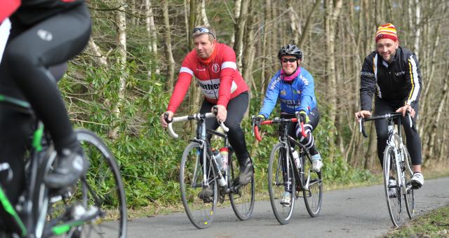 A group of people are cycling along a tarmac lane through a wood. They are all on road bikes and wearing cycling kit. They are smiling.