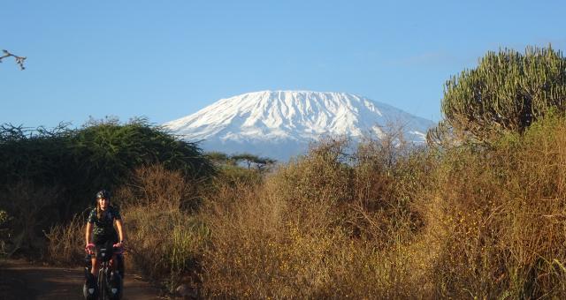 A woman on a loaded up touring bike with front and rear panniers is cycling on a dirt track through African countryside with Mount Kilimanjaro in the background