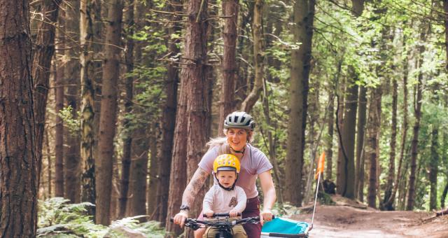 Kelly-Jayne Collinge is riding her bike on a woodland trail. Her with son Atlas is sat up front and she is pulling a trailer behind her. Mother and son are both wearing pink tops and helmets and both are smiling.