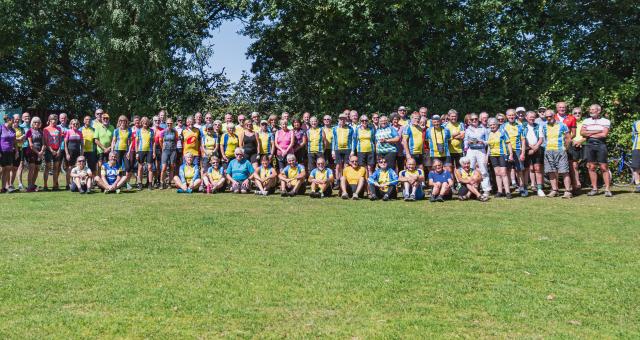 A large group of people are all gathered together on some grass with trees in the background. They are all wearing cycling clothing