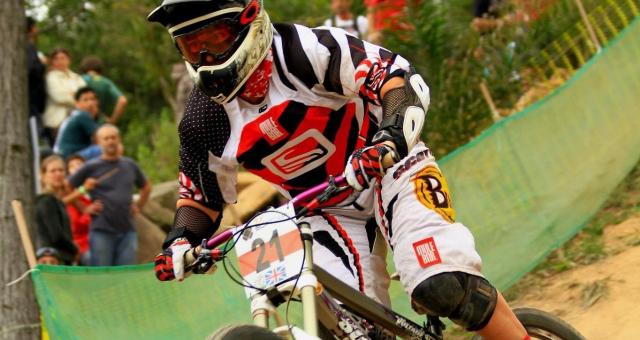 A woman is taking part in a mountain bike race. She is wearing a full-face helmet and goggles, and mountain biking clothing including knee and elbow pads. Her race number is 21 and she’s racing for Great Britain 