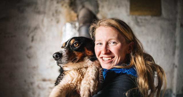 A headshot of Samantha Saskia Dugon. She has long blonde hair and is smiling into the camera. She’s holding a dog.