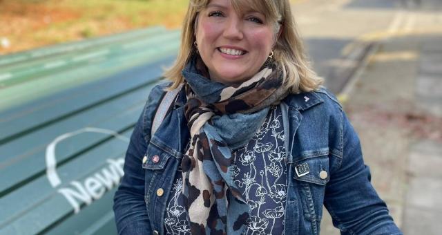 A woman is smiling into the camera. She is holding a bike and is wearing a blue denim jacket, multicoloured scarf and blue shirt with a white flower pattern. Behind her is an on-street cycle hub.