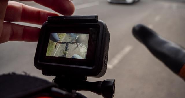 A close-up of a someone adjusting a helmet cam for a cycling helmet, the orange helmet can just be seen. The camera screen shows the bike's handlebar