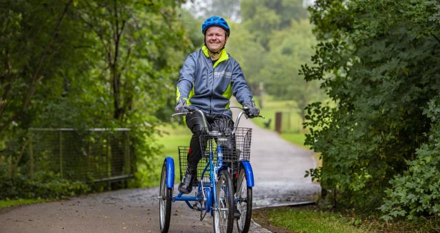 Man wearing a helmet cycling a trike outside on a footpath in a park