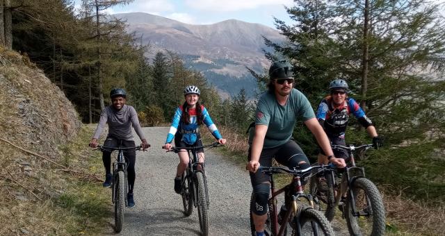 Four people are cycling along a gravel track. They're all on mountain bikes and wearing cycling kit and helmets