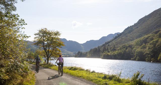 Two people are cycling on an off-road track next to a lake. They are on mountain bikes, and are both wearing shorts and T-shirts. There are mountains in the background.