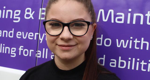 A head and shoulders shot of Sophie Bujdoso. She has long brown hair pulled back in a ponytail and is wearing glasses and a black sweater. She is smiling into the camera. She’s standing in front of the back of a van 