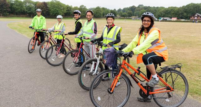 A group of people are on a tarmac path next to a field. They are all on bikes and look ready to set off on a ride wearing hi vis jackets