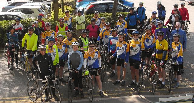 A big group of people are all standing on a pavement with their bikes. Most are wearing a Wheel Easy cycling jersey