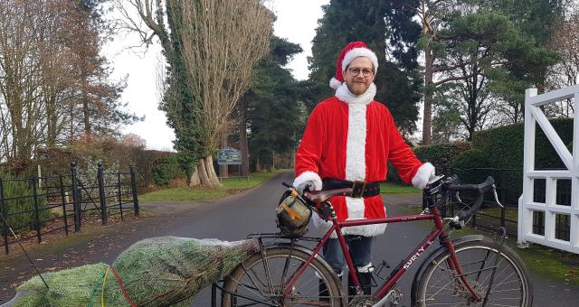 A cyclist dressed as Santa standing next to a bike with a Christmas tree in tow. 