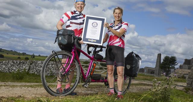 Two women in cycling kit stand either side of a pink tandem bicycle holding a framed certificate 