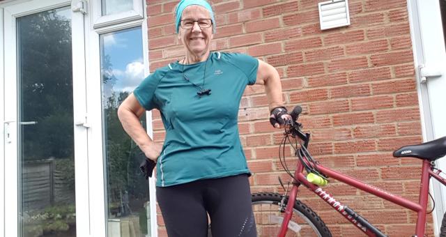 A woman wearing a azure fabric cycling top stands next to her bicycle on her patio 