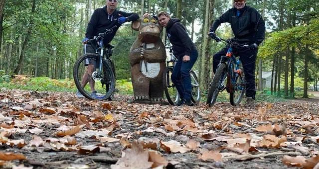 Three men on mountain bikes in a forest trail stand by a Gruffalo statue 