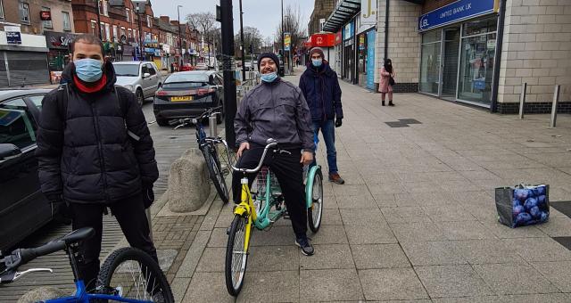 Three men spaces out and lined up along with pavement with bicycles standing close to a bus stop on a busy road