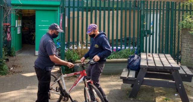 Two men stand either side of a red and white bicycle outside a green door and a set of high railings. They are standing to the left of the frame, a picnic bench is on the right. 