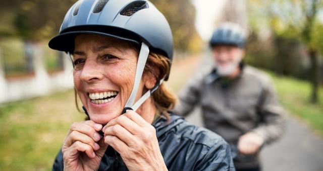 A woman adjust the chin straps of her helmet. A man can be seen out of focus in the back of the shot.