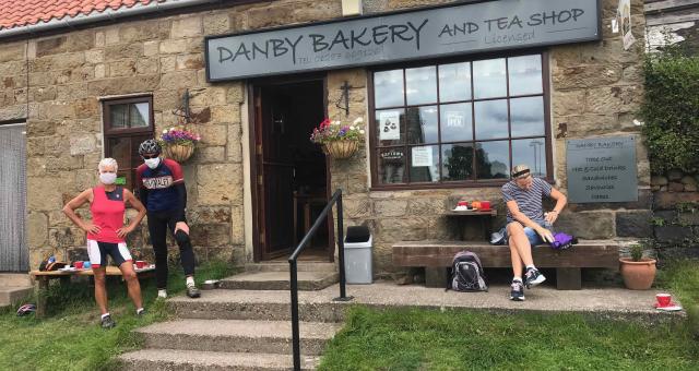 A man and woman wearing face masks pose in their cycling gear outside of a bakery. To the left of them is a lady sat on a bench tucking into her lunch