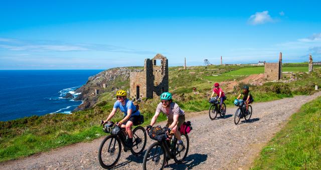 A group of cyclists ride towards camera on a gravel track along the Cornish coast. The background is scattered with tin mines