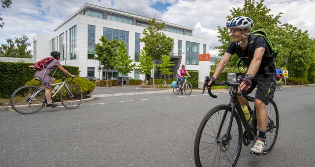 A man cycles towards camera on a road bike, he is wearing shorts and a t-shirt and carrying a backpack. In the background two women are cycling past a large office building