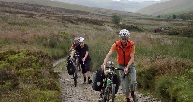 Three men on foot pushing their bikes up hill. Behind them is a beautiful lush green valley, a small abandoned building in the background