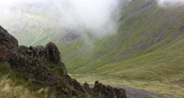 A view of Black Sail Pass, part of Wainwright’s Coast to Coast trail, showing a pass through mountains. A gravel path runs along the bottom of the pass, with grassy mountains rising up on either side. Mist is rolling in.